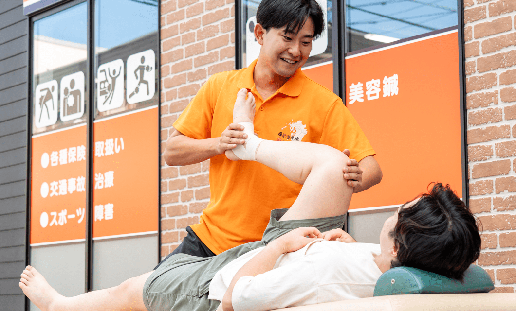 オレンジ色のポロシャツを着たセラピストが施術台で患者の脚を支え、リハビリを行っている