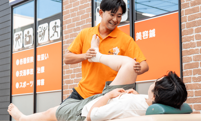 オレンジ色のシャツを着た施術者が、治療用ベッドに横たわる患者の脚を支えている場面