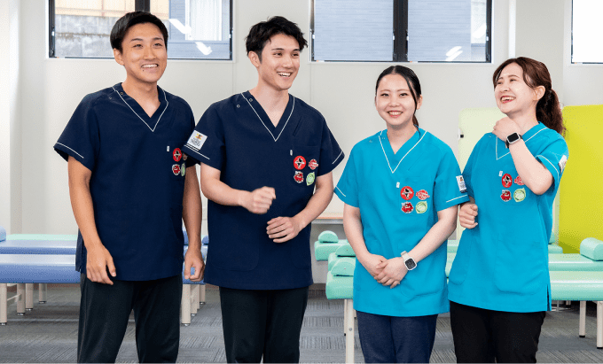 医療用スクラブを着た4人のスタッフが室内で並んで立っている。背景にはリハビリ用のベッドや椅子がある