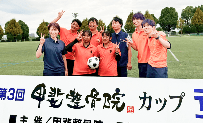 甲斐整骨院カップの看板を背景に、参加者8人が記念撮影している。1人がサッカーボールを持っている