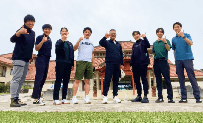 屋外で撮影された集合写真。草地の前に立つ8人が笑顔でサムズアップしている