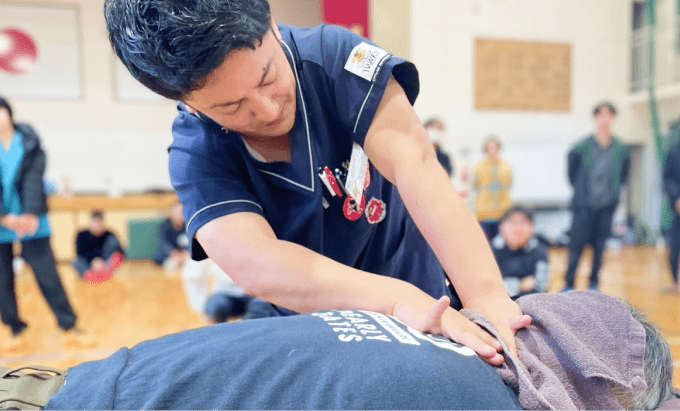 体育館のような室内で、施術者が患者の背中を押す様子を複数人が観察している