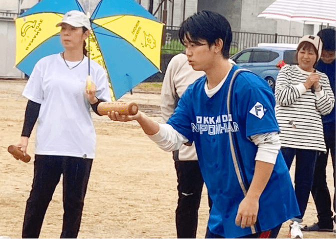 傘を持つ人々と一緒に、土の広場でモルックを楽しむ穏やかなひととき