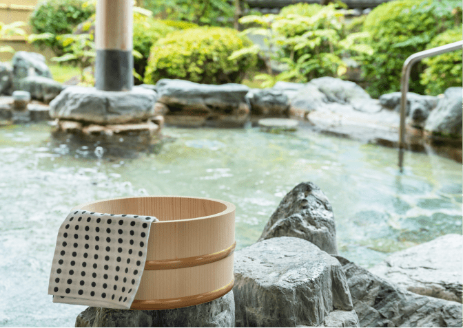 木桶と水玉模様の手ぬぐいが岩に置かれ、緑に囲まれた露天風呂の穏やかな景色