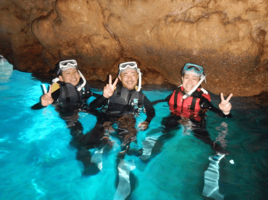 ライフジャケットとシュノーケルを着けた3人が、洞窟の青い水の中でピースサインをしている