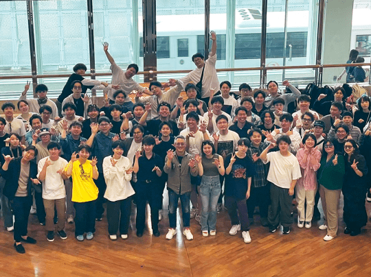 木の床の広い屋内で、大人数が笑顔やジェスチャーを交えて楽しそうに集合写真