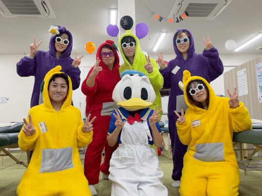 パーティー風の室内で、カラフルな仮装をした7人が笑顔で記念撮影