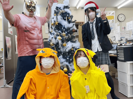 オフィス風の室内で、クリスマスツリーを背景にキャラクター衣装や仮装をした仲間たちが楽しげに記念撮影