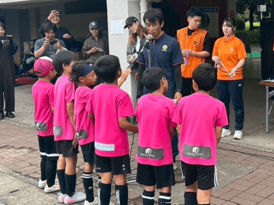 スポーツイベントの表彰式で、子どもたちがユニフォーム姿で並び、大人がマイクで称える場面